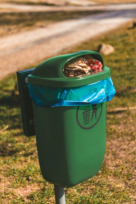 A weathered red metal waste container with visible rust and chipped paint is situated against a dark green, textured wall on the left and a light gray, rough concrete wall on the right. The container 